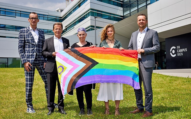 © HCW/Ludwig Schedl Gruppenfoto mit fünf Personen, die die Regenbogenfahne auf der Wiese vor dem Haupteingang der F H Campus Wien halten