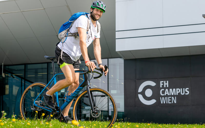 Studierender steht auf Fahrrad vor der Hochschule Campus Wien
