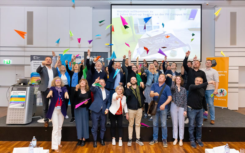 (c) FH Campus Wien/Schedl Gruppe von Menschen auf einer Bühne wirft bunte Papierflieger