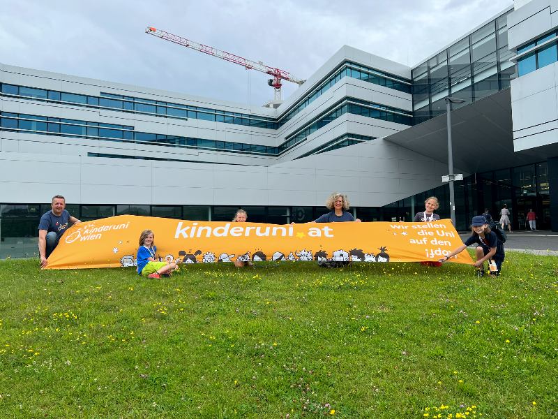 Zwei Erwachsene und drei Kinder stehen vor einem Gebäude und halten ein Banner hoch