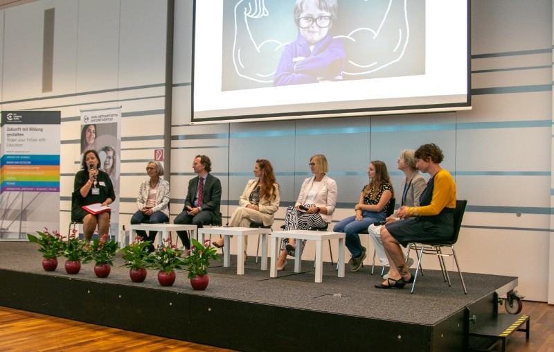 Personen sitzen auf einem Podium, im Hintergrund ist eine Leinwand zu sehen