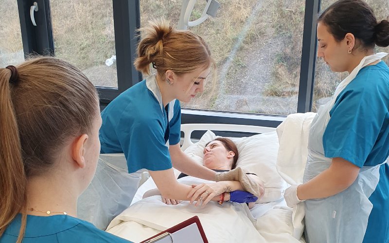 Studierende an einem Krankenbett mit einer Schauspielpatientin über die Körperpflege