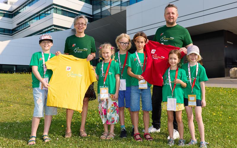 Eine Gruppe von Personen, darunter eine erwachsene Frau, ein erwachsener Mann und sechs Kinder stehen vor einem Gebäude in einer Wiese. Sie tragen T-Shirts auf denen Kinderuni geschrieben steht.