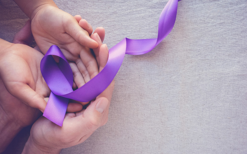 (c) istock/SewcreamStudio Hände einer erwachsenen Person und eines Kindes halten eine Lavendelschleife als Zeichen für Solidarität bei Krebserkrankungen