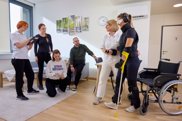 © HCW/Schedl In einem hellen Therapieraum üben mehrere Personen gemeinsam das Gehen. Eine Patientin mit Augenbinde und Trainingsausrüstung wird dabei von einer Therapeutin begleitet. Zwei weitere Therapeutinnen sowie drei Personen beobachten und unterstützen die Übung. Neben der Patientin stehen ein Gehstock und ein Rollstuhl.