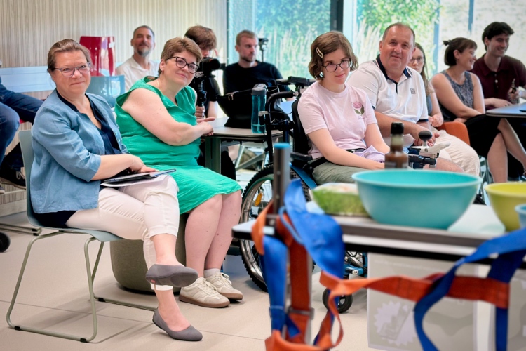 © HCW Mehrere Personen sitzen in einem hellen Seminarraum und verfolgen aufmerksam eine Präsentation. Im Vordergrund steht ein Tisch mit Schüsseln und bunten Hebegurten, im Publikum sitzt unter anderem eine Person im Rollstuhl. Die Teilnehmenden blicken nach vorne und wirken interessiert.