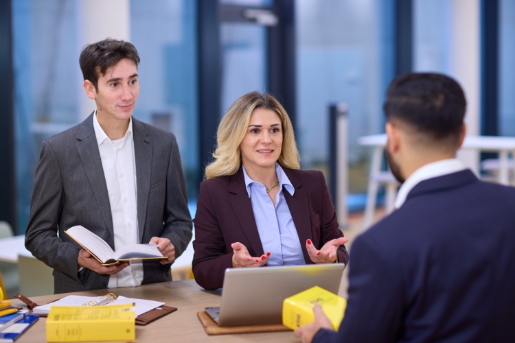 © HCW/Schedl Drei Personen stehen an einem Tisch. Auf dem Tisch liegen Bücher und ein Laptop.