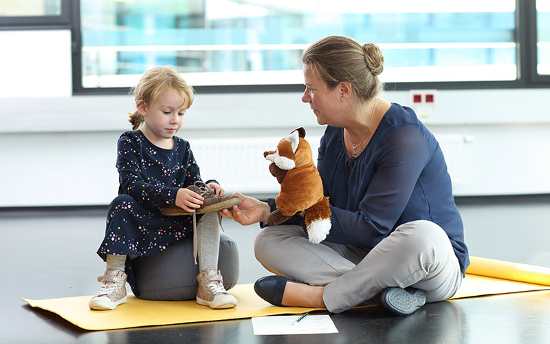 © HCW / Schedl Ergotherapeutin sitzt mit Kind am Boden und sie üben gemeinsam das Schuhbänder binden.