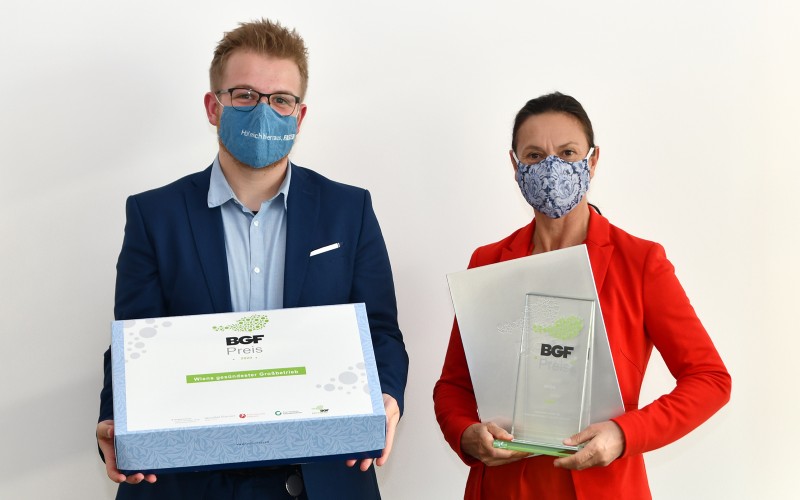 Das Campus Vital-Team, Raphael Hengl und Susanne Mulzheim, stehen von einer weißen Wand und halten den BGF-Preis und eine Geschenkbox. 