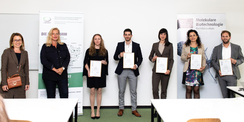 Sieben Personen stehen vor zwei Roll-ups und einer Wand, fünf Personen halten ihre Urkunden der Preisverleihung in die Kamera