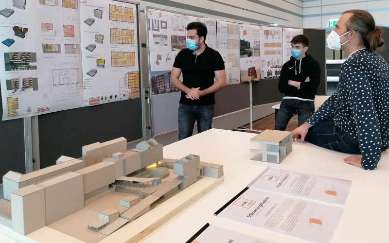 zwei männliche Studierende vor Flipcharts, präsentieren dem Juror Pläne, auf einem Tisch stehen Architekturmodelle