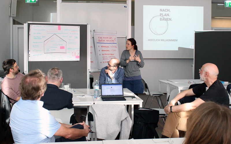 Vortragende*r in einem Seminarraum mit einer Gruppe Zuhörenden an Tischen sitzend, im Hintergrund: Flippcharts, Tafel und darauf das Logo Nach.Plan.Bauen.