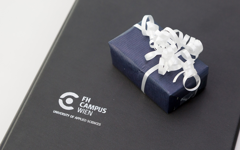 © HCW Schwarze Urkundenmappe mit dem Logo der Hochschule Campus Wien und kleinem Geschenk in dunkelblauem Papier und mit weißemBand