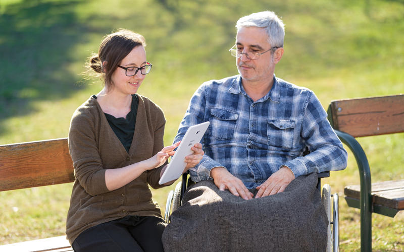 (c) FH Campus Wien / Schedl Im Park ein älterer Mann in einem Rollstuhl, daneben eine jüngere Frau, sie beide beschäftigen sich mit einem Tablet