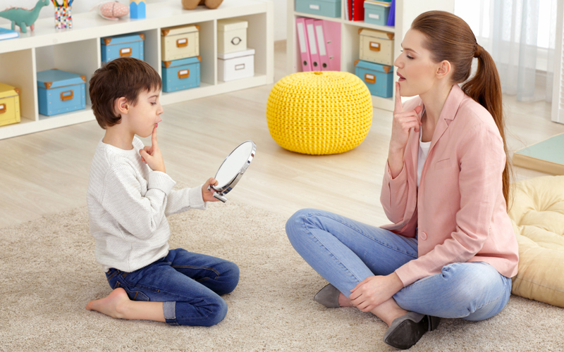 © Africa Studio / Shutterstock.com Eine Logopädin sitzt mit einem Jungen auf dem Boden. Er hat einen Spiegel vor sich und macht gemeinsam mit der Logopädin eine Sprechübung