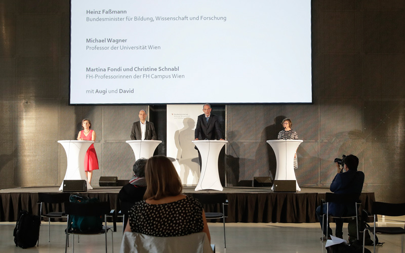 Vier Personen stehen in Abständen von 1,5 Metern auf einem Podium an weißen Stehtischen für die Pressekonferenz. Links außen steht Martina Fondi von der Hochschule Campus Wien, dann Michael Wagner von der Universität Wien, es folgt Heinz Faßmann, Bundesminister für Bildung und Christine Schnabl von der Hochschule Campus Wien. Hinter ihnen sind ihre Namen eingeblendet, davor sitzen in Abständen die Vertreter*innen der Medien auf Stühlen