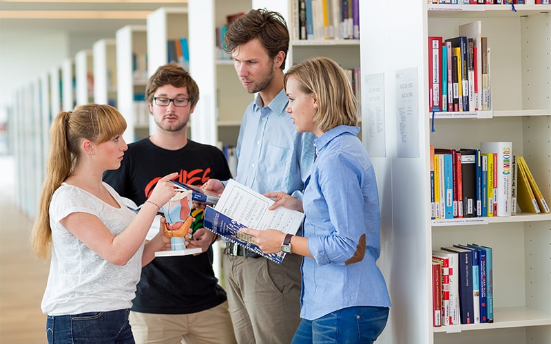 © HCW/Ludwig Schedl Vier Studierende stehen in einer Bibliothek und unterhalten sich