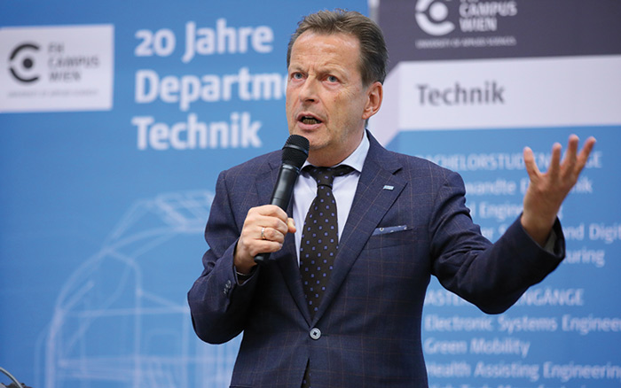 © Schedl / FH Campus Wien Wilhelm Behensky spricht zu einem Publikum und hält ein Mikrofon in seiner Hand während die andere gestikuliert