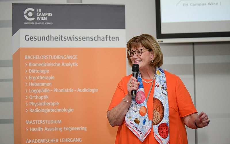 Dame in oranger Bluse mit Mikrofon in der Hand vor Gesundheitswissenschaften Rollup der Hochschule Campus Wien