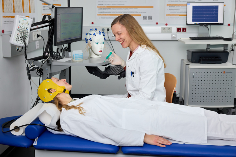 Eine Person mit EEG-Haube auf einer Untersuchungsliege. Daneben eine Person im Kittel vor einem Monitor, die die Untersuchung durchführt. 