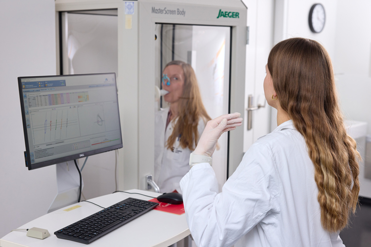 Eine Person im Laborkittel vor einem Monitor, die Anweisungen an eine Patientin in einer Untersuchungskammer gibt.