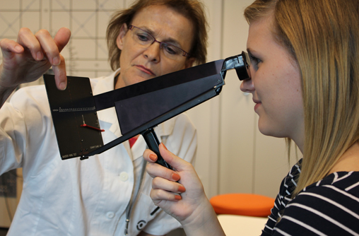 Studierende hält ein schwarzes Instrument in der Hand, in der Form von einer Brillenfassung ohne Bügel. Vor der Brille befindet sich eine dunkele kleine Skala Karte, welche mit der Brille verbunden ist. Sie hält das Instrument beim Griff in der Mitte des Geräts und schaut durch Form in Brillenfassung. Die Lehrende steht neben der Skala Karte und zeigt mit dem Finger auf einen Punkt.