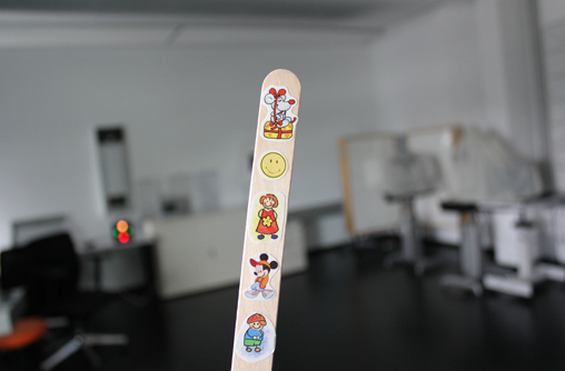 Ein länglicher Holzstab auf den kleinen Figuren untereinander abgebildet sind. Die erste Figur ist eine Cartoon Maus auf einem Stück Käse. Die zweite Figur ist ein gelbes, rundes, Smiley Gesicht das lächelt. Die dritte Figur ist ein gezeichnetes Mädchen mit rotem Kleid. Die vierte Figur ist die Disney Cartoon Figur Mickey Mause. Die letzte Figur ist ein gezeichneter Junge mit einem blauen T-Shirt und einer grünen Hose.