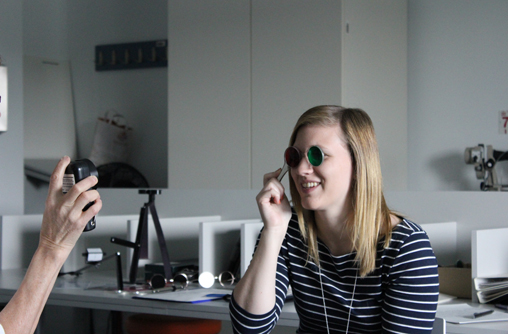 Die Studierende hält ein Instrument in der Hand, besteht aus einem Glasvorhalter, in der Form von Brillenfassung ohne Bügel, dafür mit einem seitlichen Griff. Gegenüber ist ein Teil der Hand der Vortragenden zu sehen, welche ein kleines schwarzes rechteckiges Gerät in der Hand hält und auf das Gesicht der Studierenden richtet.