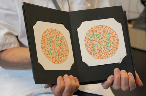 Bildausschnitt von den Händen einer Person, mit weißen Kittel, die ein offenes Buch zur Prüfung des Farbensinnes oder Farbensehens hochhält. Auf den Seiten sind kleine, unterschiedlich große und grünfarbige Punkte zu sehen, die eine Zahl bilden. Der Hintergrund ist ebenfalls mit unterschiedlich große und rot und orangefarbigen Punkte bedeckt.