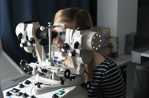 Die Studierende blickt in den Synoptophor. Sie stützt ihr Kinn auf eine dafür vorgesehene Fläche, auf dem Gerät ab und hat links und rechts, vor Ihren Augen, eine röhrenförmig Form zum reinblicken. An dem Gerät sind viele Schrauben und Knöpfe, seitlich sind Lichter die aufleuchten.