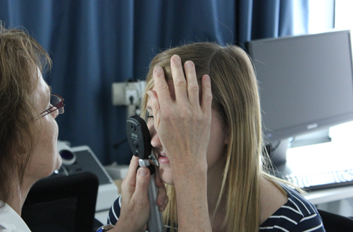 Studierende wie sie gegenüber der Lehrende sitzt. Diese hat ein kleines, schwarzes, handliches Untersuchungsgerät in der Hand. Das Gerät besteht auf einem Griff und drauf befindet sich eine runden, dicken Scheibe, in der Größe eines Auges. Die Lehrende hält in der linken Hand das Gerät, vor dem rechten Auge der Studierende. Die linke Hand legt Sie auf die Stirn der Studierende, um sie zu positionieren.