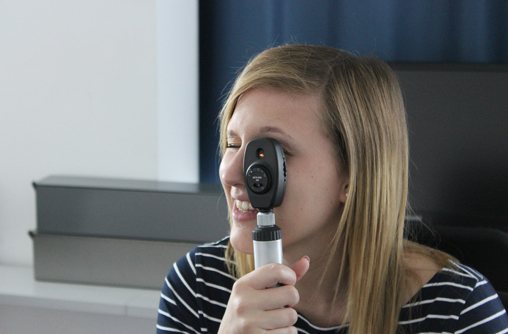 Lächelnd blickt die Studierende, mit ihrem linken Auge, durch das handliches, schwarzes, und kleines Untersuchungsgerät. Das rechte Auge hat sich dabei geschlossen.