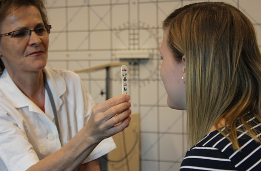 Die Studierende sitzt gegenüber einer Lehrenden die ein kleines Holzstäbchen auf Augenhöhe hält. Es sind sechs bunte Formen oder Figuren auf den Stab aufgedruckt, wie zum Beispiel ein Elefant, ein Teddybär. Die meisten Formen und Figuren sind nicht erkennbar, da sie verschwommen fotografiert sind.