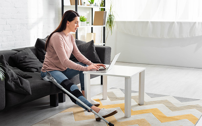 Eine Frau mit Beinschiene sitzt auf einer Couch und arbeitet an einem Laptop.