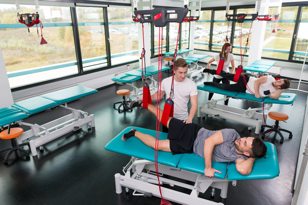 Studierende liegen seitlich auf einer Liege im Funktionsraum. Ihre rechten Füße sind in einer Schlaufe, welche von einem Gerät an der Decke hängt, somit hängt der Fuß in der Luft. Am Ende der Liegen stehen andere Studierende, die Übungen mit den liegenden Studierende durchführen. Dabei berühren Sie die Füße der Liegenden Personen und bewegen diese.