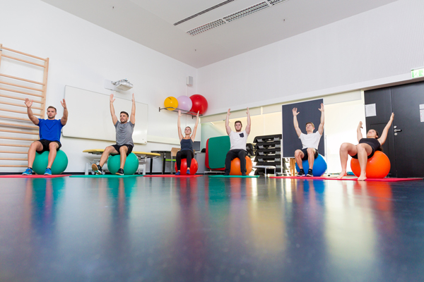 Studierende sitzen auf bunten Gymnastikbällen im Kreis und machen dabei eine Übung, bei der sie die Hände ausgestreckt in die Höhe halten.