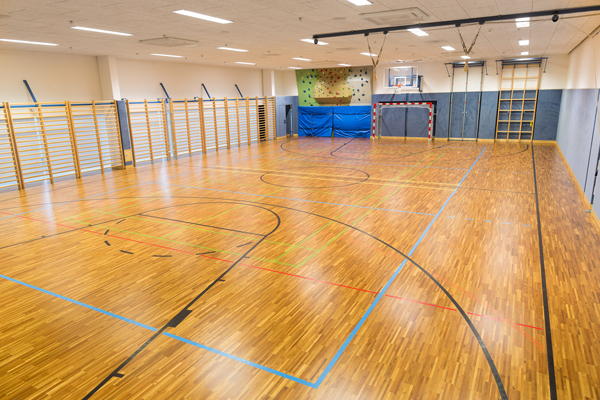 Leerer Turnsaal. Links im Raum befinden sich Sprossenwände, vorne im Raum ist eine Kletterwand daneben ein Fußballtor.