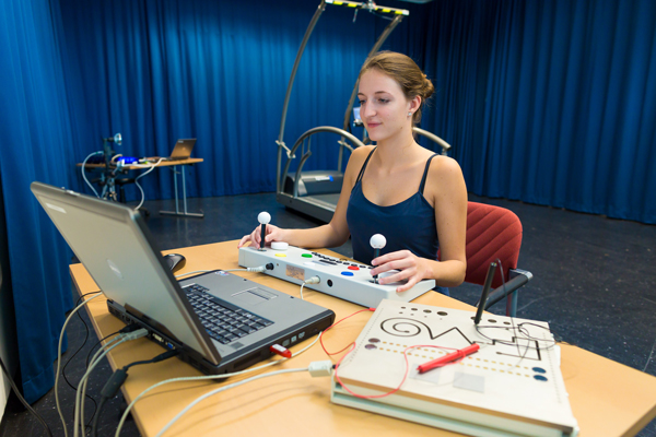 Studierende sitzt vor einem Laptop an dem eine Art Kontroller angeschlossen ist, mit vielen Knöpfe und zwei Steuerknüppel. Sie schaut auf den Bildschirm vom Laptop und bewegt dabei die Steuerknüppel.