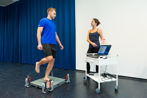 Studierende steht mit einem Bein auf eine Fußmessplatte, den anderen Fuß hält er hinten in die Höhe. Gegenüber von ihm ist ein Computer, auf einen Rollwagen, der Druckverteilung des Fußes grafisch darstellt. Neben dem Computer ist eine andere Studierende, die auf die Grafik zeigt und sich mit dem Studierenden unterhält.