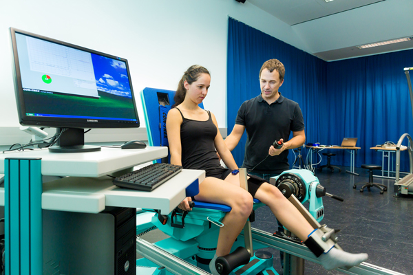 Eine Studierende sitzt in einem Sessel. Ihr linker Fuß ist dabei an eine längliche Installation vom Sessel angeschlossen, die den Widerstand bei Kniestreckung misst. Sie hebt den linken Fuß. Neben ihr steht ein Mann mit einem Knopf in der Hand. Der Knopf ist mit einem Kabel verbunden, welches zum Sessel führt. Er blickt dabei das Knie der Studierende an.