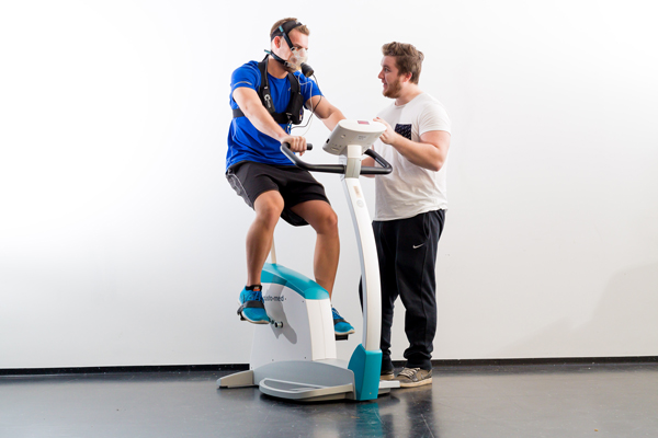 Ein Studierender sitzt auf einem Ergometer mit Atemmaske und tritt in den Pedalen, neben ihm steht ein anderer Studierende und unterhält sich mit ihm. Das Ergometer hat die Form eines Fahrrades ohne Räder. Beide blicken einander an.