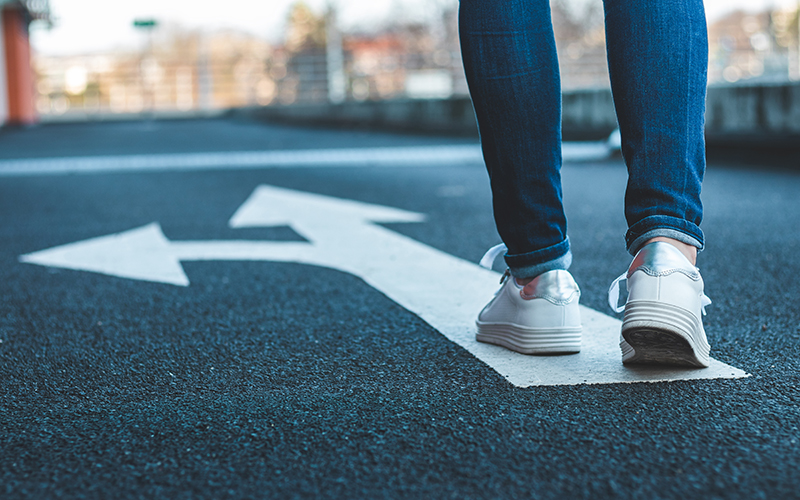 © encierro/shutterstock Eine junge Person folgt einer Straße, auf dem Teer sind weiße Bodenmarkierungen. Ein Pfeil zeigt geradeaus, der andere biegt nach Links ab.