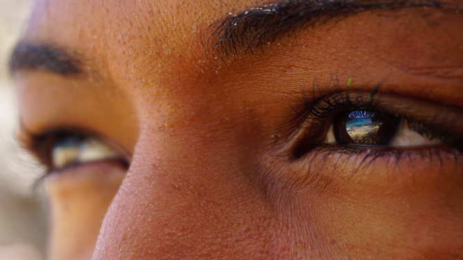 Nahaufnahme von Augen einer jungen Frau, in den Augen spiegelt sich der Strand