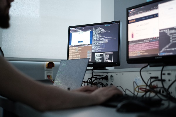 Person sitzt vor mehreren Bildschirmen und arbeitet an Tastatur. Düstere Bildstimmung