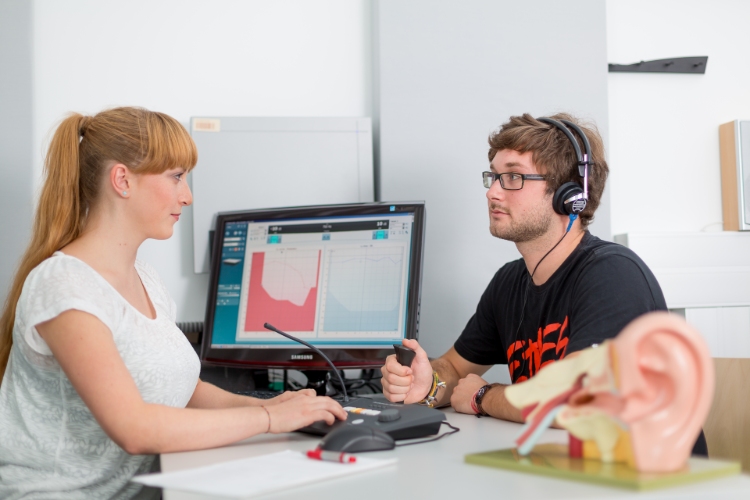 Zwei Personen sitzen gegenüber an einem Tisch. Hinten steht ein Bildschirm, der Mann recht trägt Kopfhörer.