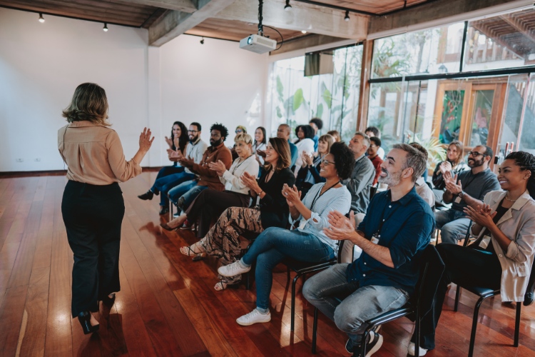 Eine Gruppe von Menschen sitzt in einem modernen großen Raum. Vorne steht eine Frau und präsentiert.