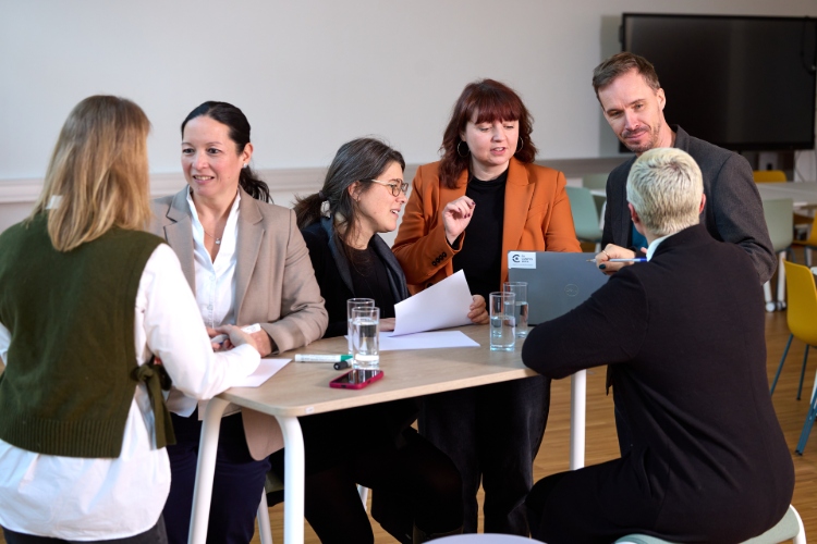 Eine Gruppe von Erwachsenen sitzt an einem Tisch in einem hellen Seminarraum und bespricht Unterlagen. Auf dem Tisch liegen Ausdrucke, ein Laptop und Gläser mit Wasser; die Personen sind im Gespräch und gestikulieren, als Teil einer gemeinsamen Arbeits- oder Lernsituation.