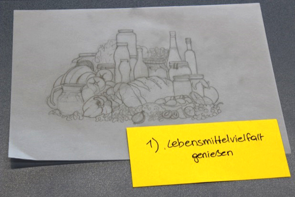 Ein Blattpapier auf dem Lebensmittel wie Brot, Paprika, Weintrauben, Wasserkrug, unterschiedlich große Flaschen, Zwiebeln, Zitrusfrüchten und vielen mehr, mit Bleistift gezeichneten ist. Auf dem Bild liegt ein weiterer gelbfarbiger Zettel, mit der Aufschrift Lebensmittelvielfalt genießen