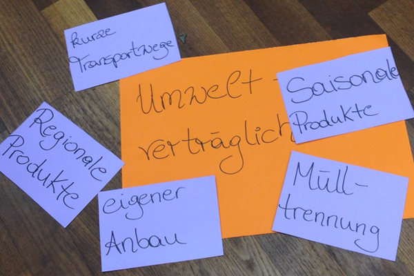 Bildausschnitt von einem Tisch, auf dem sich ein orangefarbiger Zettel mit der Aufschrift Umwelt verträglich befindet. Rund um diesen Zettel sind kleiner violettfarbene Zettel, mit den Aufschriften Regional Produkte, eigener Anbau, kurze Transportwege, Saison Produkte und Mülltrennung