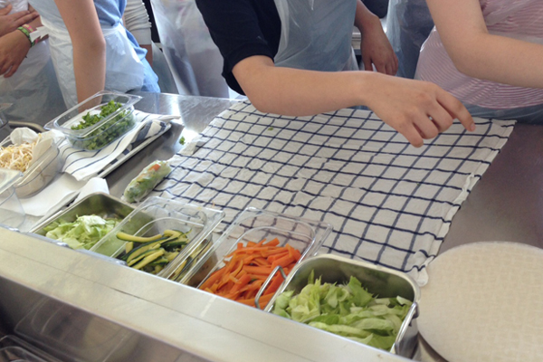 Mehrere Personen stehen vor einem Tisch, auf den sich unterschiedliche Schüssel mit klein geschnitten Gemüse und Kräutern befindet, wie zum Beispiel Karotten, Salat, Sojasprossen und vieles mehr.